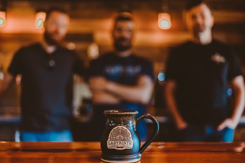 In focus picture on a black beer mug with clay logo medallion with blurred background of three men standing side by side.